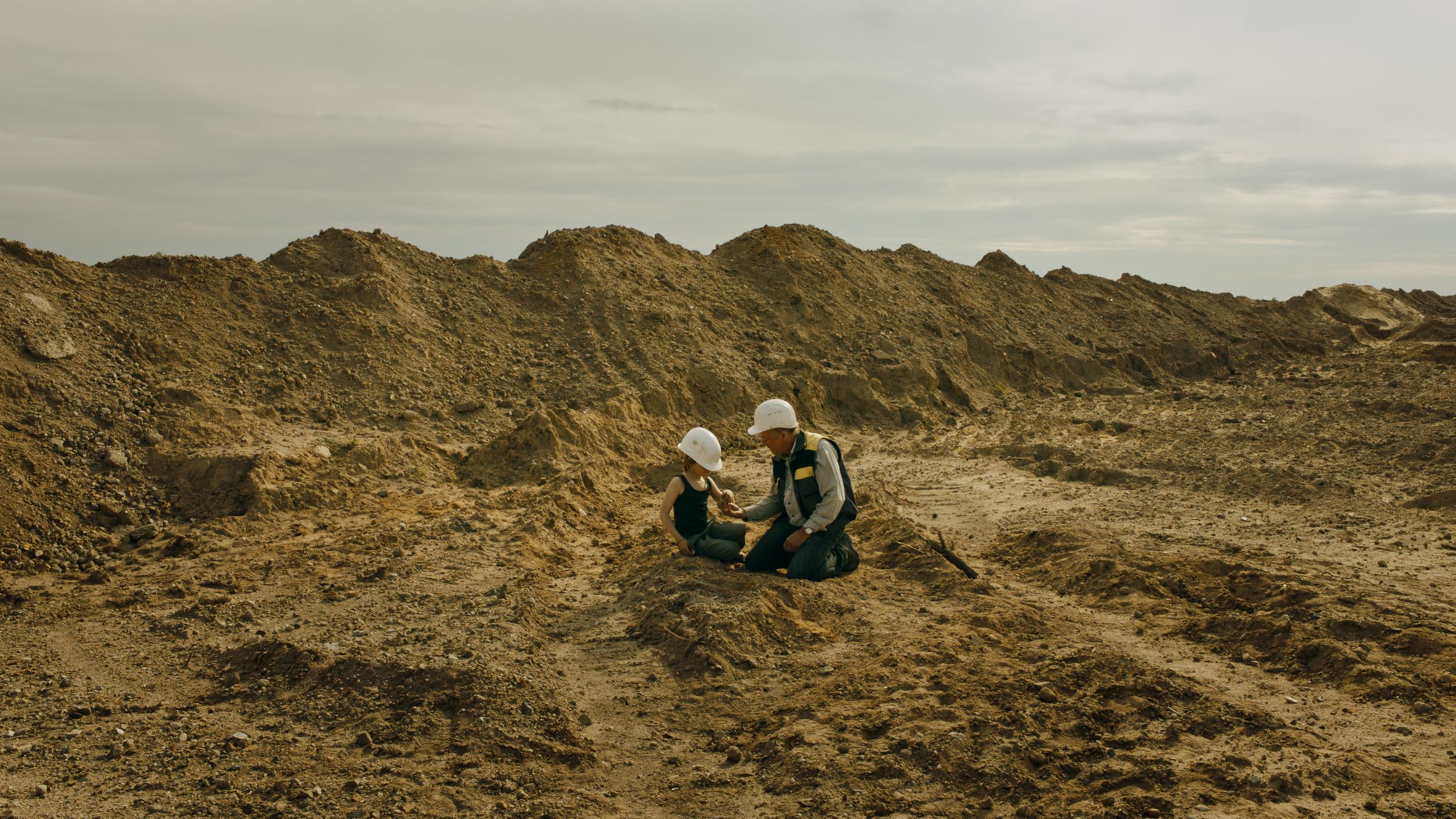 "Fossil": Michael (Markus Hering) und Toni (August Schulz) sitzen inmitten einer Landschaft aus Sand und Erde. Beide tragen weiße Schutzhelme und betrachten, was Toni in der Hand hält.