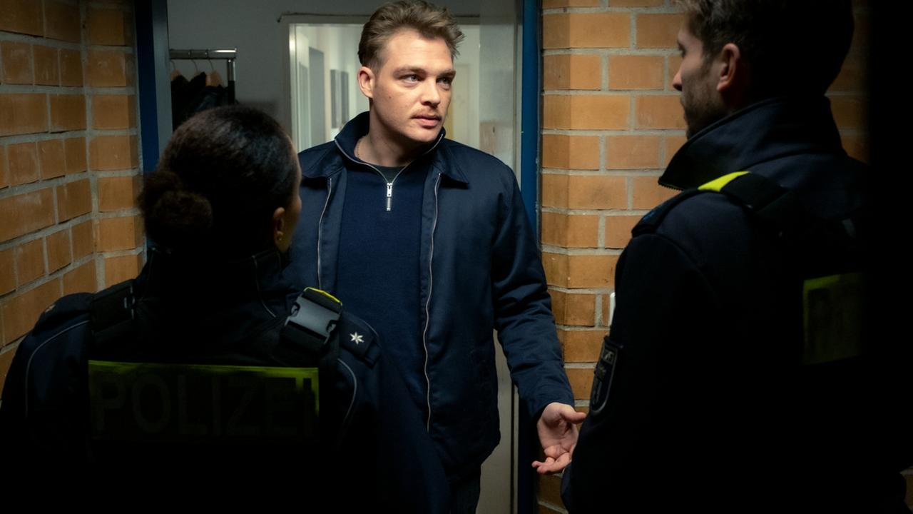 "Wahrheit oder Lüge - Die Macht der Frauen": Polizist Leon (David Schüttler) in Zivilkleidung steht in einem Gebäudeflur und unterhält sich  mit zwei Polizeibeamten in Uniform
