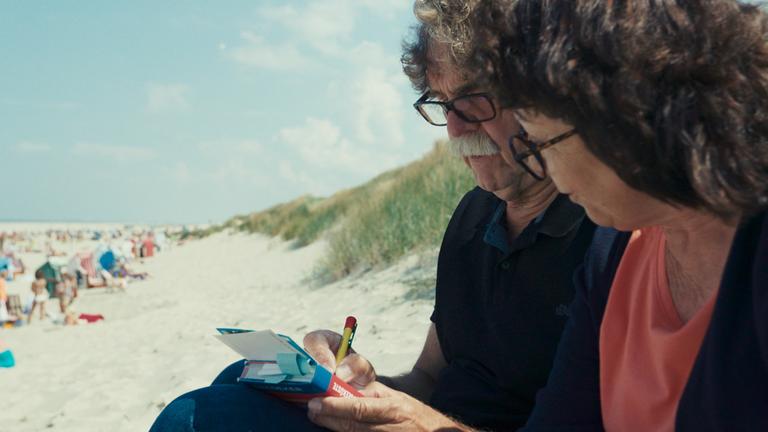 Ein älteres Ehepaar sitzt im Sand am Dünenrand eines Strandes. Der Mann schreibt Postkarten; die Frau hält ein Buch als Schreibunterlage