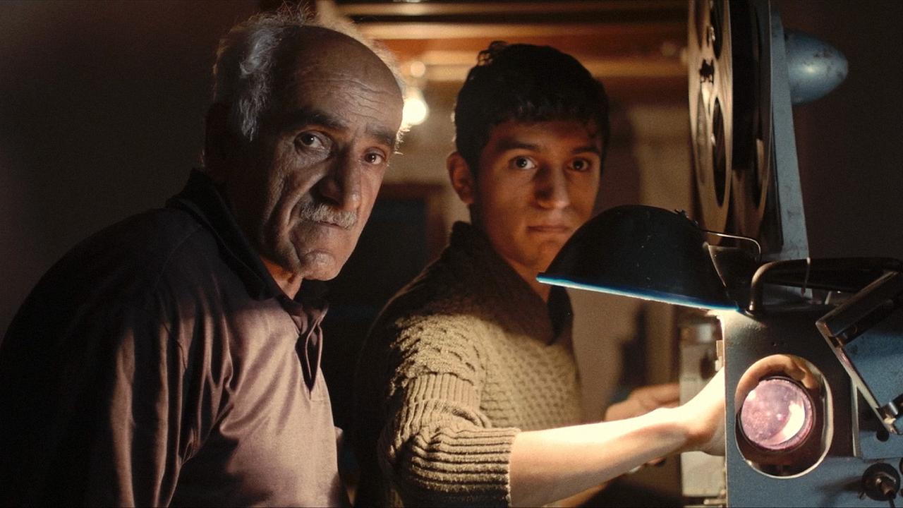 "Die Rückkehr des Filmvorführers": Ein älterer und ein junger Mann stehen im Vorführraum eines altertümlichen Kinos an einem alten Filmprojektor und blicken in die Kamera