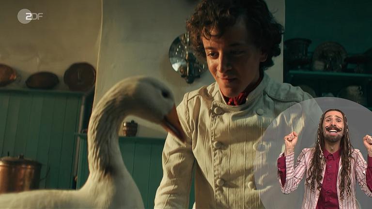 Screenshot "Zwerg Nase":  Zwerg Nase (Mick Mehnert). Im Vordergrund steht eine Gans. Rechts  unten ist der Gebärdendolmetscher in einem Kostüm eingeblendet 