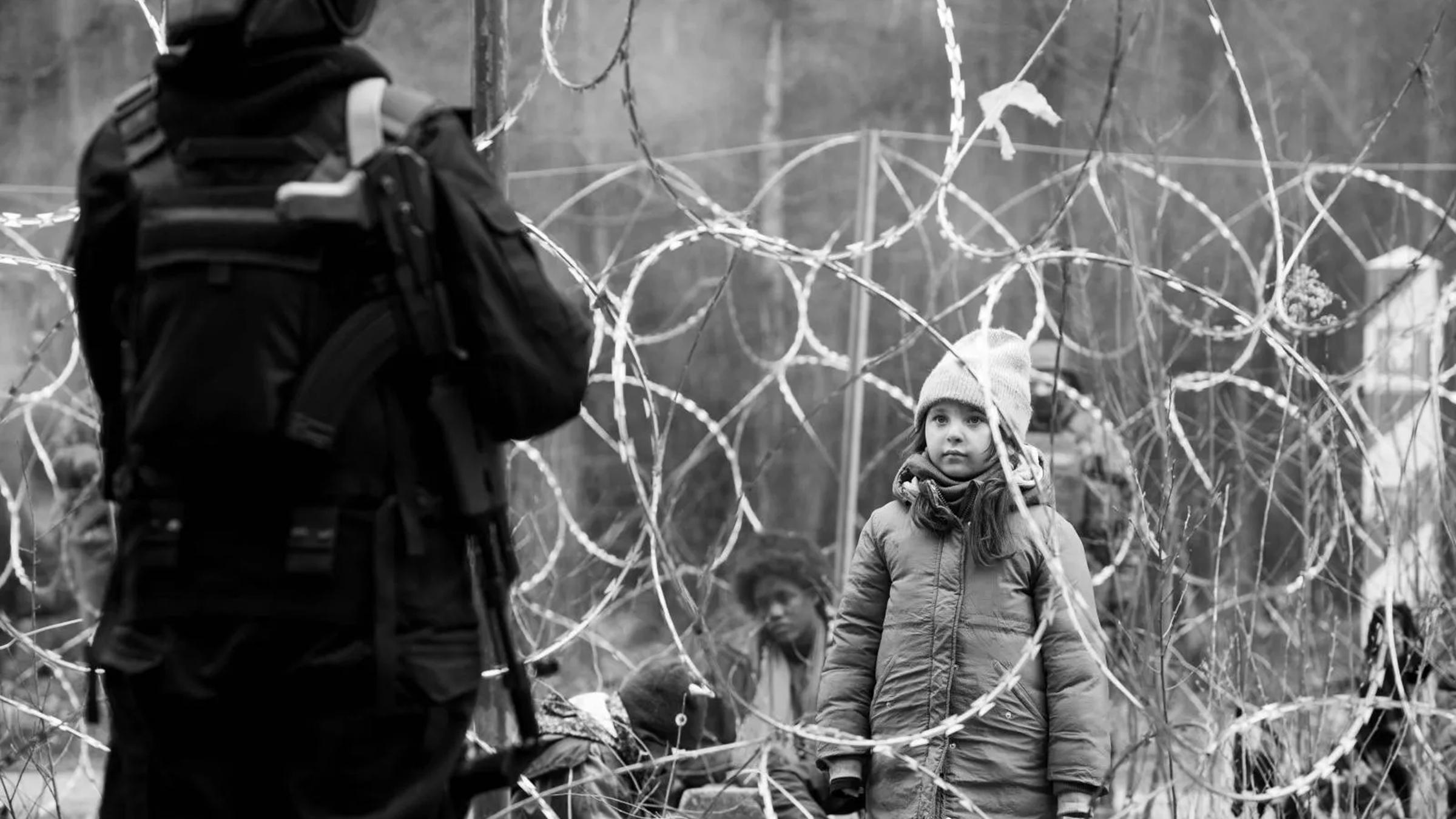 "Green Border": Ein kleines Mädchen steht vor einem undurchdringlichen Stacheldrahtzaun. Im Vordergrund mit dem Rücken zur Kamera ein Grenzsoldat. Im Bildhintergrund ein Grenzpfahl und weitere Migranten.