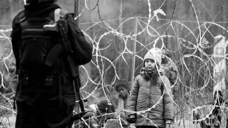 "Green Border": Ein kleines Mädchen steht vor einem undurchdringlichen Stacheldrahtzaun. Im Vordergrund mit dem Rücken zur Kamera ein Grenzsoldat. Im Bildhintergrund ein Grenzpfahl und weitere Migranten.