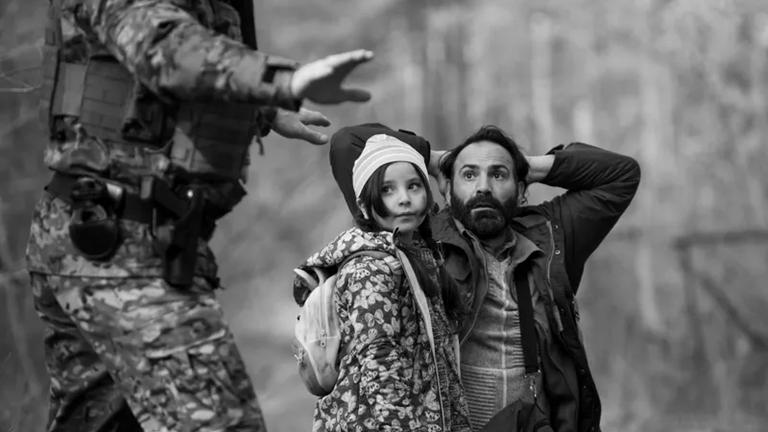 "Green Border" (Filmszene): Ein kleines Mädchen drängt sich an einen knienden Mann, der seine Arme hinter dem Kopf verschränkt und angsterfüllt zu einem Soldaten links im Bildvordergrund blickt.