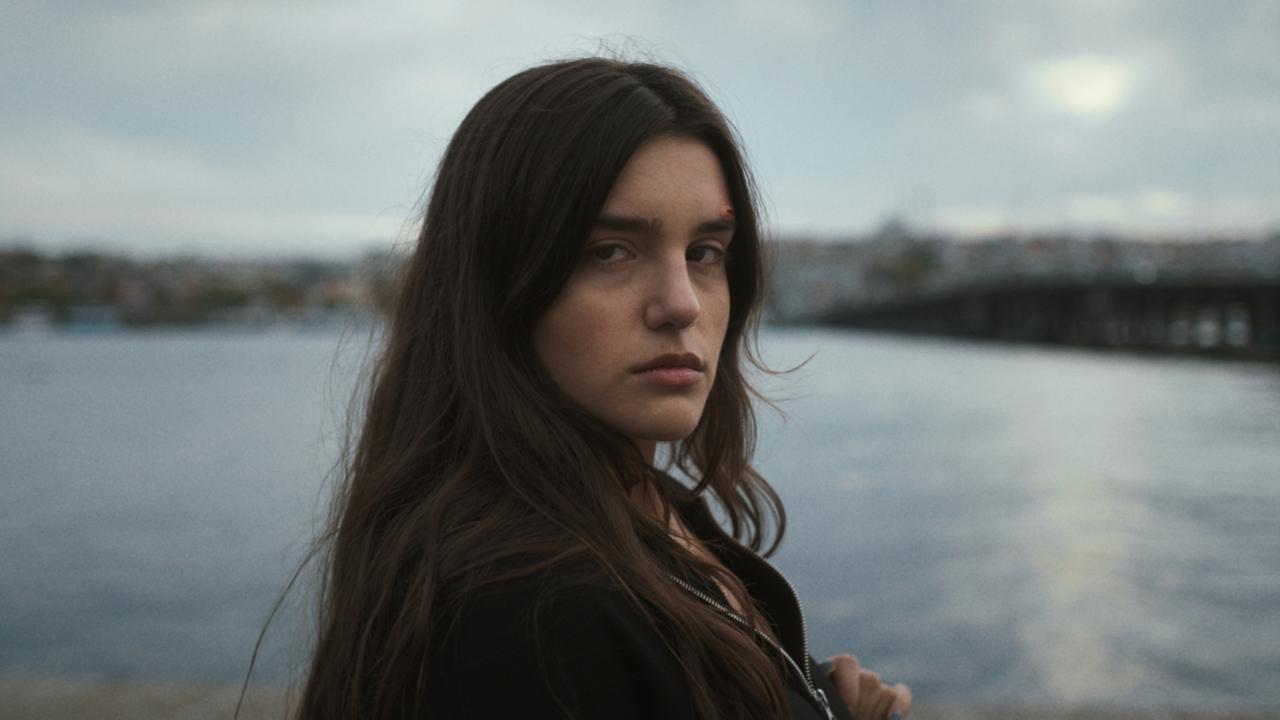 Hazal (Melia Kara) steht an einem See und blickt mit einem verunsiocherten Gesichtsausdruck in die Kamera.