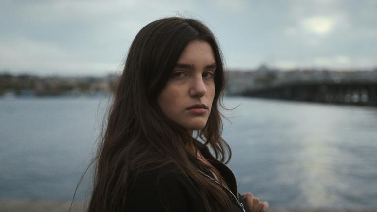Hazal (Melia Kara) steht an einem See und blickt mit einem verunsiocherten Gesichtsausdruck in die Kamera.