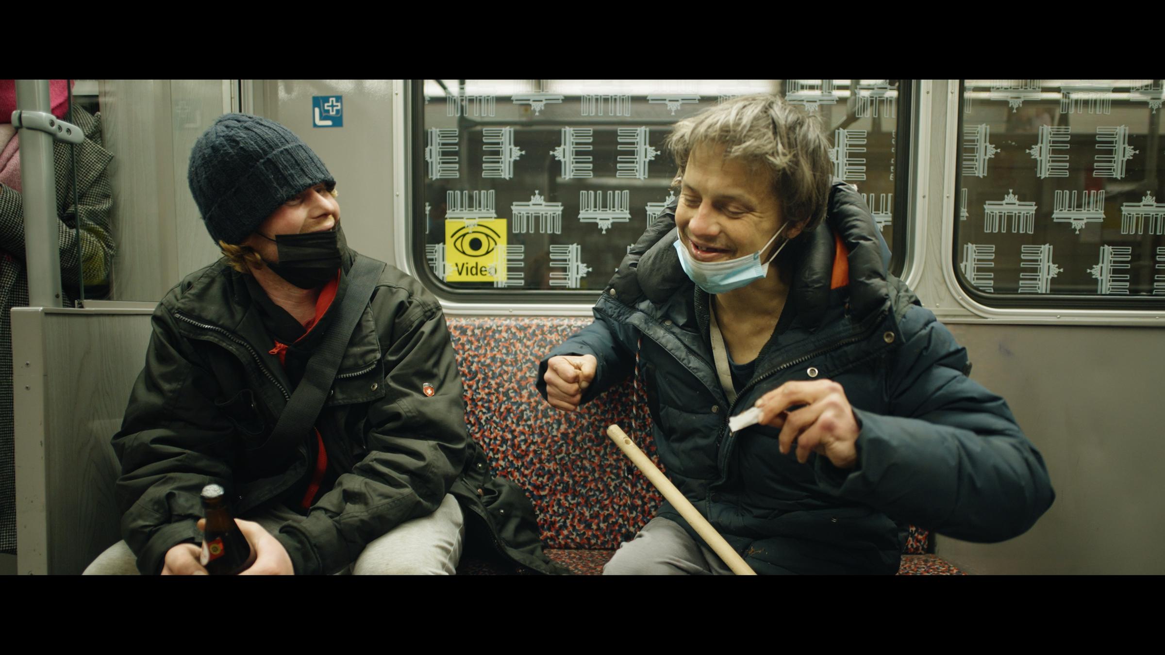 "Hausnummer Null": Eine Frau und ein älterer Mann sitzen in der S-Bahn nebeneinander und unterhalten sich. Die Frau trägt eine Mütze und eine FFP 2-Maske und umklammert mit beiden Händen eine Bierflasche. Der Mann hat seine Maske aufs Kinn runtergezogen und dreht sich eine TZigarette. Auf seinem Schoß ein Besenstiel.