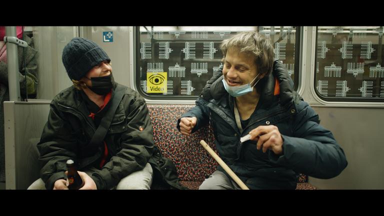 "Hausnummer Null": Eine Frau und ein älterer Mann sitzen in der S-Bahn nebeneinander und unterhalten sich. Die Frau trägt eine Mütze und eine FFP 2-Maske und umklammert mit beiden Händen eine Bierflasche. Der Mann hat seine Maske aufs Kinn runtergezogen und dreht sich eine TZigarette. Auf seinem Schoß ein Besenstiel.