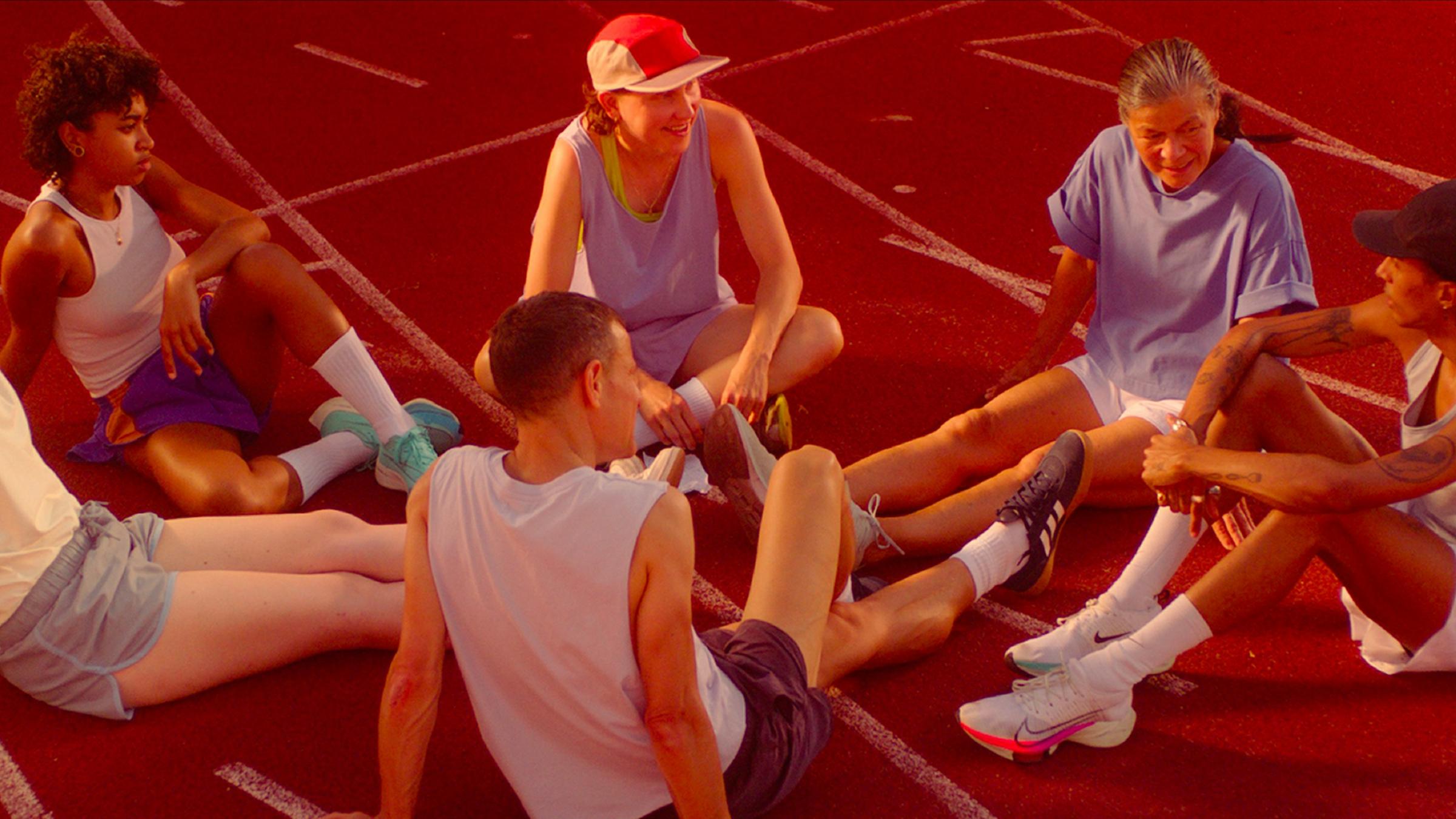 Queer gewinnt - Eine Sport-Utopie ("Life is not a competition, but i'm winning"): Athlet*innen eines queer-feministischen Kollektivs im Leichtathletik-Dress sitzen entspannt auf einer Tartanbahn und unterhalten sich