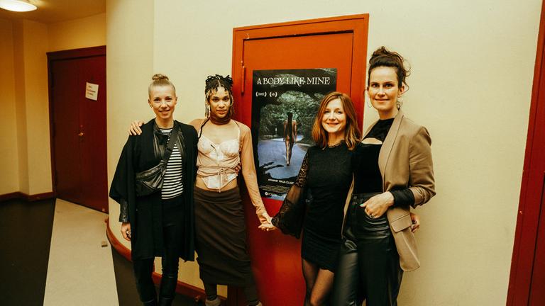 "Ab 18! - A Body like mine": v.l.n.r.: Alina Albrecht, Puck, Maja Classen und Saralisa Volm stehen voreinem an einer roten Tür angebrachten Filmplakat und lächeln in die Kamera. 