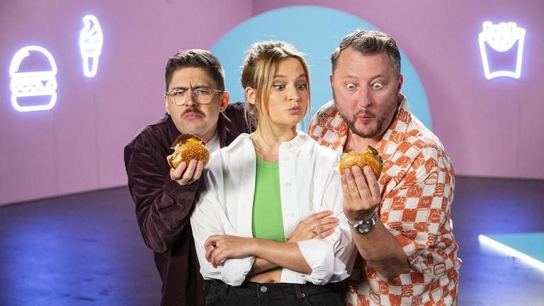"besseresser: Die miese McPlant-Show": Sebastian Lege (r.) und und Florian Reza (l.) stehen im Studio nd blicken verwundert und skeptisch auf die Burger, die sie in ihren Händen halte. Zwischen Ihnen steht Lilly Temme mit verschränken Armen und blickt pikiert auf den Buirger in Sebastian Leges Hand. 