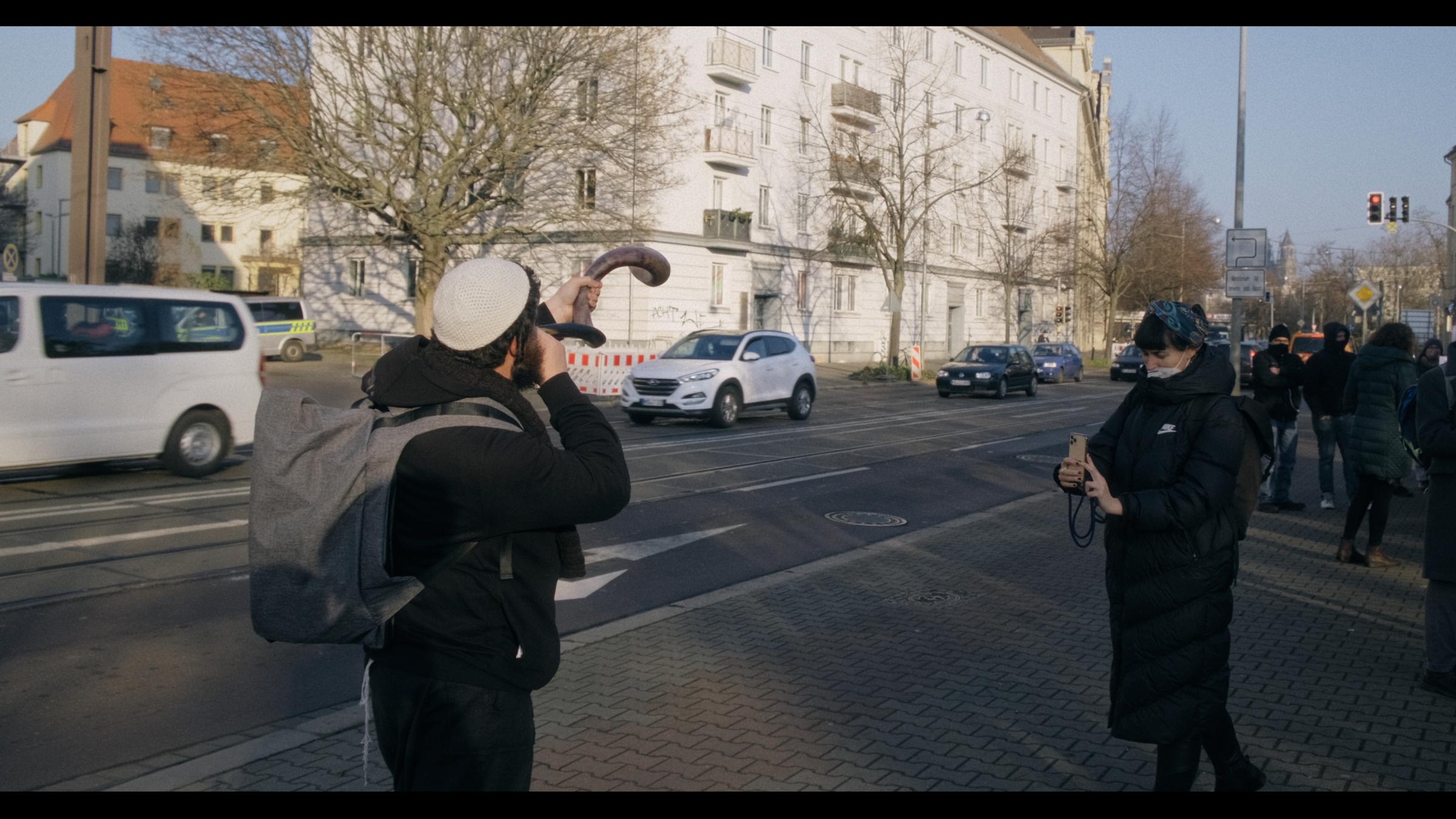 "Einzeltäter - Halle": Vor dem Gerichtsgebäude in Halle befinden sich mehrere Personen. Im Vordergrund bläst ein jüdischer Gläubiger mit einer "Kippa" bekleidet in ein traditionelles jüdisches Horn, er wird von einer Frau mit einem Smartphone gefilmt.