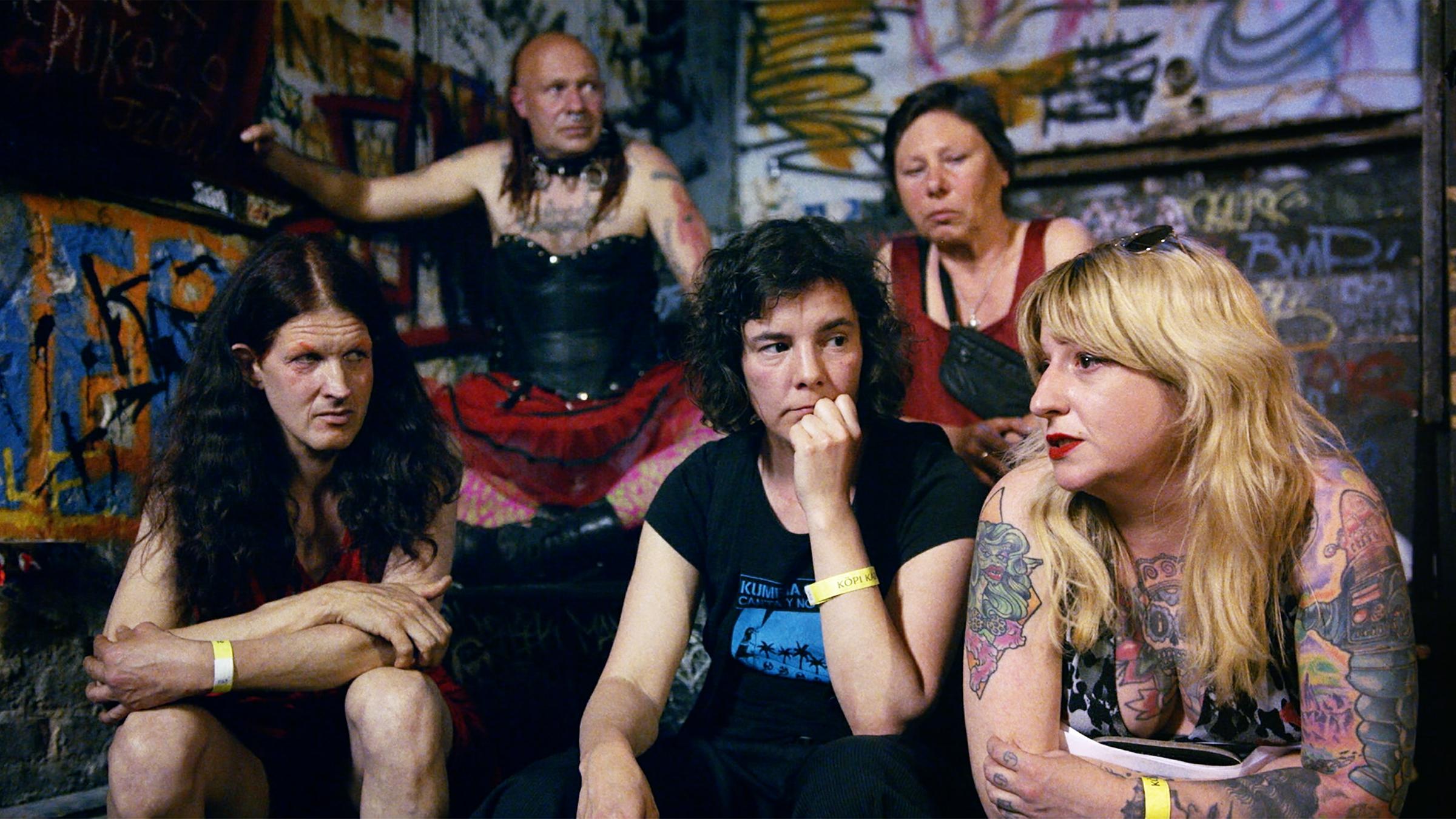 "Berlin Utopiekadaver": Zwei Männer und fünf Frauen sitzen vor einer Graffittiwand und blicken nachdenklich.