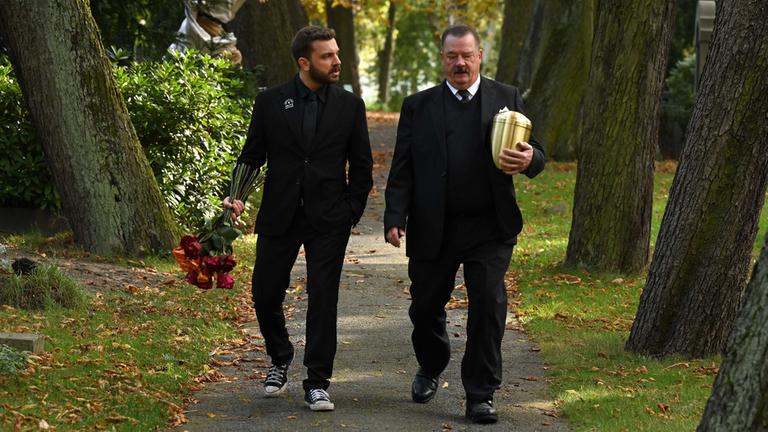 "Sterben für Beginner": Eric (Edin Hasanovic, l.) und Bestatter Volker Mutz (Peter Kurth, r.) laufen nebeineinander auf einem baumbestandenen Friedhofsweg und unterhalten sich dabei. Beide tragen schwarze Trauerkleidung. Eric hat einen großen Strauß Rosen in der rechten Hand; Volker Volker rägt in der linken Armbeuge eine goldene Urne.