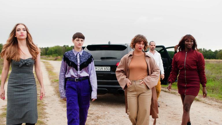 Die geschützten Männer:  Sina Martens, Johanna Polley, Britta Hammelstein und Ruby Commey (v.l.n.r.) laufen nebeneinander auf einem Feldweg, dahinter läut Yousef Sweid. Hinter ihnen steht ein schwarzes Auto mit offenenTüren 