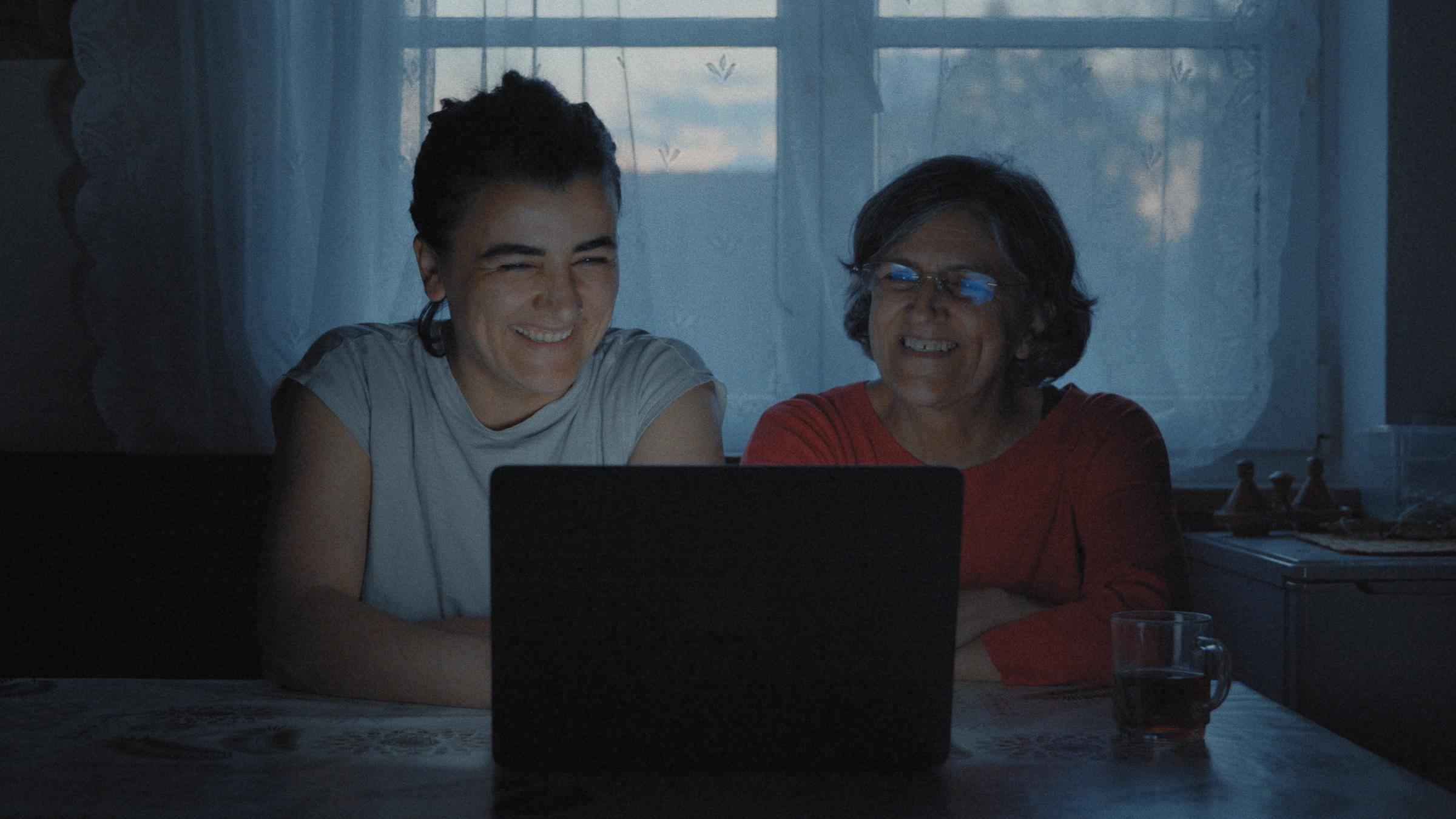 "Exile Never Ends": Mutter Yildiz Bektaş und Tochter Bahar Bektaş schauen lachend auf einen Laptop, der auf einem Küchentisch vor ihnen steht.