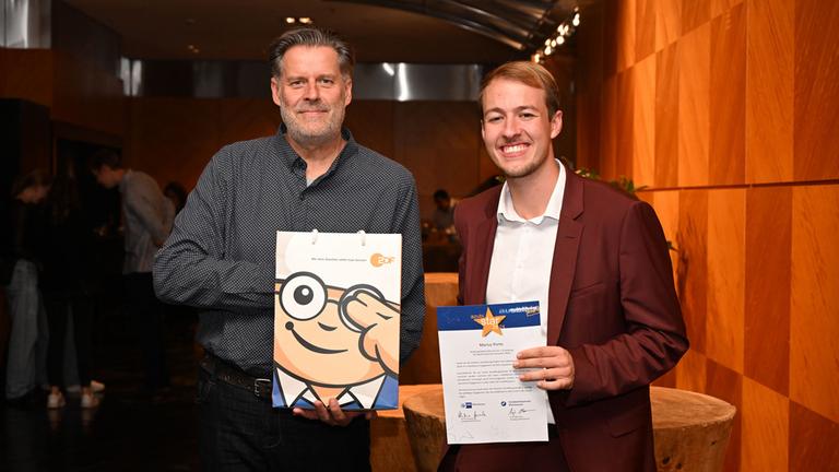 IHK Rheinhessen - Ehrung der zwölf besten Auszubildenden aus der Region im Hyatt Regency Mainz, 6. September 2024: Marius Porte, Azubi-Star 2024, präsentier die Auszeichnung. Neben ihm hält sein sein Ausbilder Stephan Ullrich eine Mainzelmännchen-Tüte in die Kamera. Beide lächeln