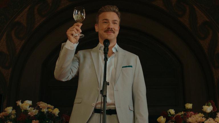 "Pfau - Bin ich echt?": Matthias (Albrecht Schuch) steht lächelnd vor einem Mikrofon und prostet mit einem Weinglas in der rechten Hand dem Publikum zu.