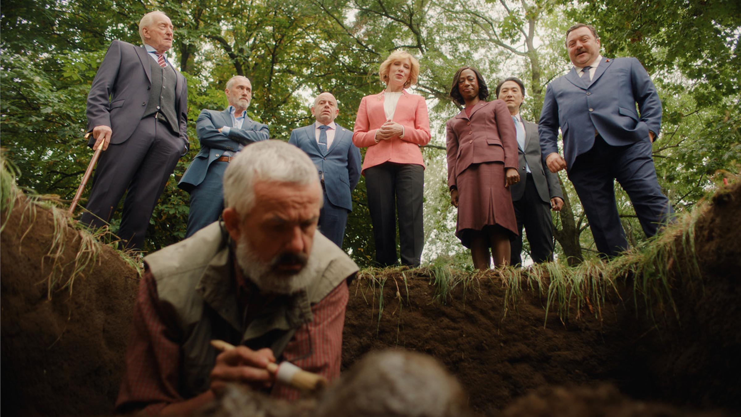 "Rumours": Hilda Orlmann (Cate Blanchett, M.), Edison Wolcott (Charles Dance, l.), Maxime Laplace (Roy Dupuis, 2. v. l.), Antonio Lamorte (Rolando Ravello, 3. v. l.), Cardosa Dewindt (Nikki Amuka-Bird, 3. v. r.), Tatsuro Iwasaki (Takehiro Hira, 2. v. r.) und Sylvain Broulez (Denis Ménochet, r.) stehen vor einer Ausgrabungsstelle im Wald und beobachten die Arbeit eines Anthropologen (Ralph Berkin, vorne), der mit einem Pinsel eine Leiche säubert