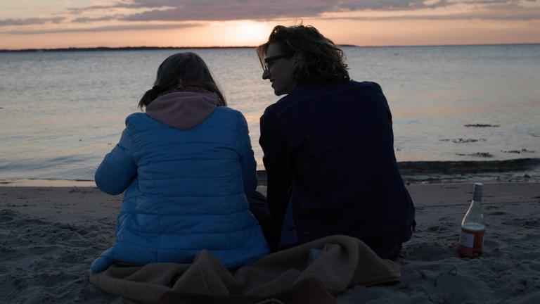 "Demenz: Mensch sein, Mensch bleiben" ("Human Forever") (Arte): Altenpfleger Teun Tobes sitzt mit dem Rücken zur Kamera bei Sonnenuntergang auf einer Decke am Strand und blickt dabei eine weibliche Person an an, die links neben ihm sitzt. Links neben ihm im Sand eine halbleere Weinflasche; im Hintergrund geht die Sonne über dem Meer unter.