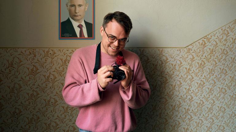 "Mr. Nobody against Putin":  Pavel Talankin steht vor einer Wand und lächelt in dier Kamera. Er trägt ein rosa Sweatshirt und hält mit beiden Händen einen Fotoapparat fest, den eer an einem Gurt um seinen Hals trägt und an dem eine rote Blume steckt. Im Hintergrund an der Wand ein Porträt von Wladimir Putin.