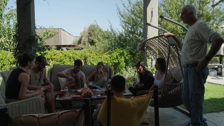 "Hamas–Angriff aufs Festival – Die Überlebenden des Wüsten-Raves": Einge Gruppe von sieben Jungen Leuten sitzt auf einer schattigen Veranda um einen Tisch herum. Sie trinken Kaffee und unterhalten sich. auf der echten Seite steht ein älterer Mann und blickt in die Runde.