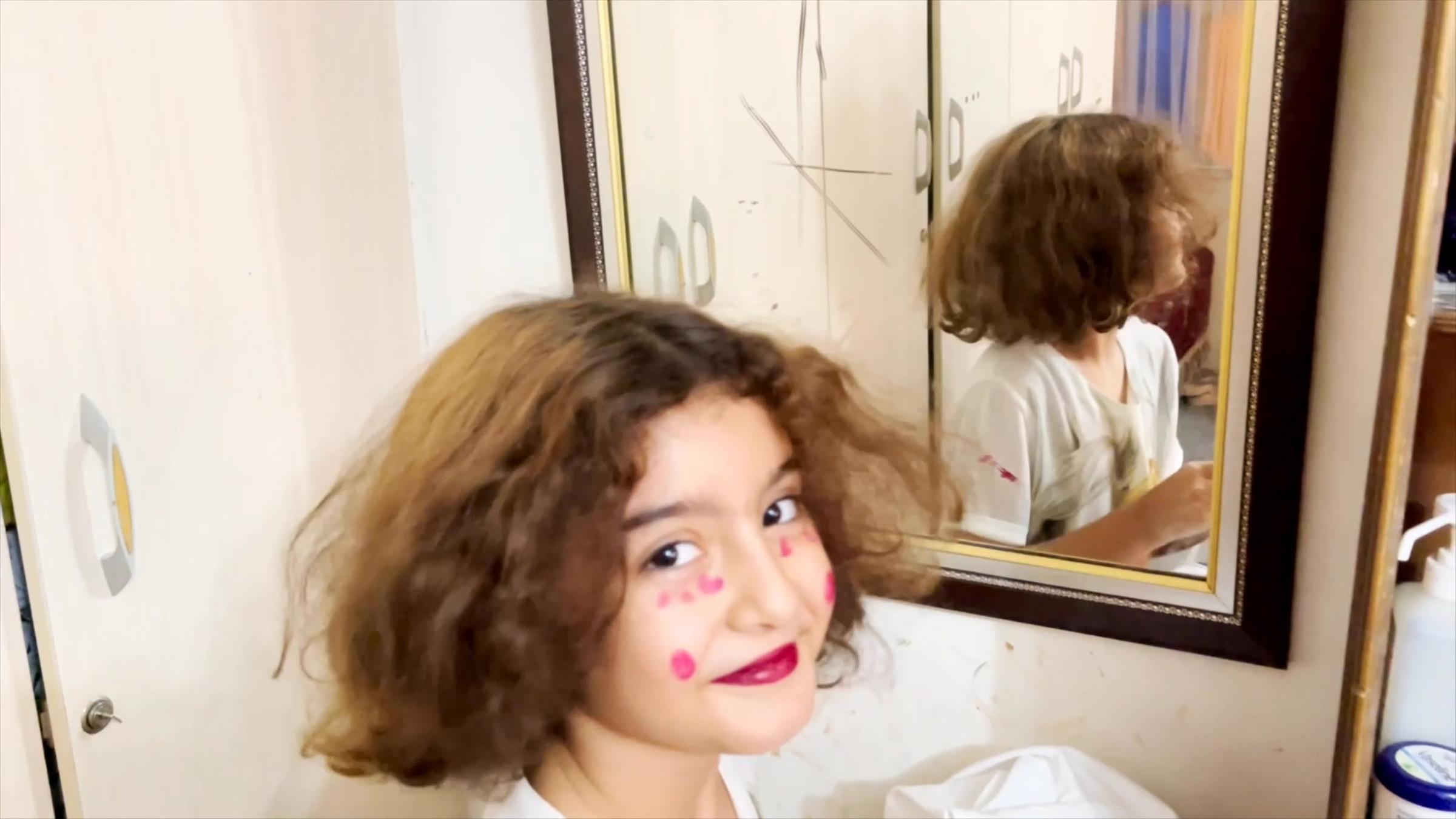 "Nilas Traum vom Garten Eden": Nila sitzt zwischen Kleiderschrank und Spiegel und lächelt in die Kamera. Sie hat sich mit Lippenstift und roten Punkten im Gesicht geschminkt.