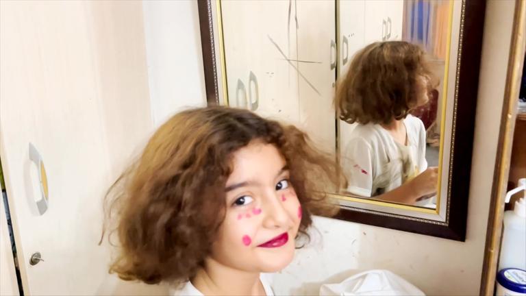 "Nilas Traum vom Garten Eden": Nila sitzt zwischen Kleiderschrank und Spiegel und lächelt in die Kamera. Sie hat sich mit Lippenstift und roten Punkten im Gesicht geschminkt.