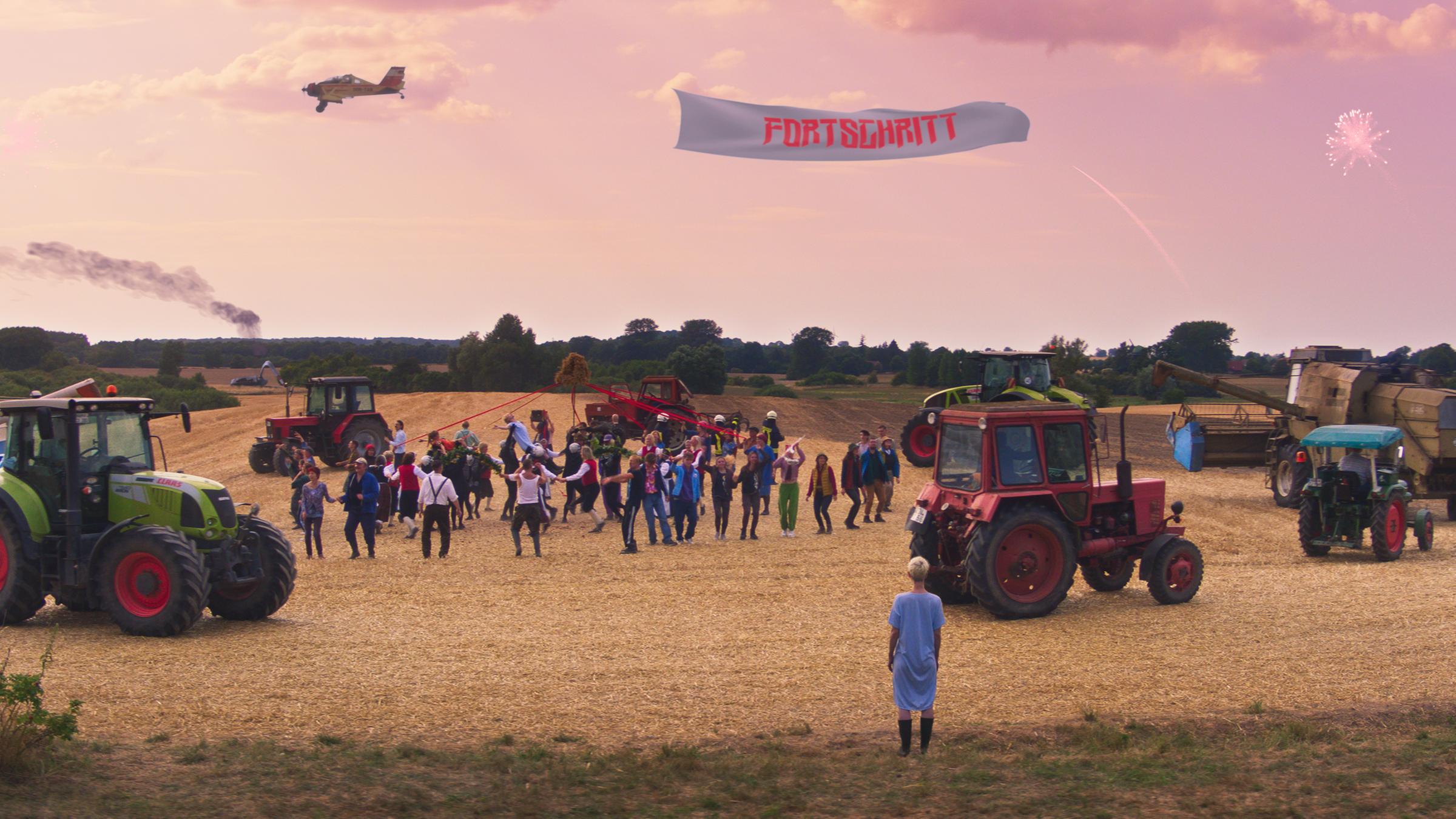 "Rote Sterne überm Feld": Auf einem abgeernteten Feld tanzt eine Gruppe von 20 - 30 Menschen, kreisförmig umringt von Traktoren und Mähdreschern. Darüber fliegt ein Flugzeug, das ein Banner mit der Aufschrift "Fortschritt" hinter sich herzieht.