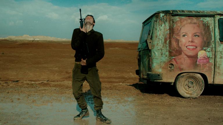 FOXTROT: Ein Mann steht mit erhobenem Kopf und geschlossenen Augen in der Wüste und umklammert dabei ein Gewehr. Rechts im Bild ein heruntergekommener Lieferwagen.