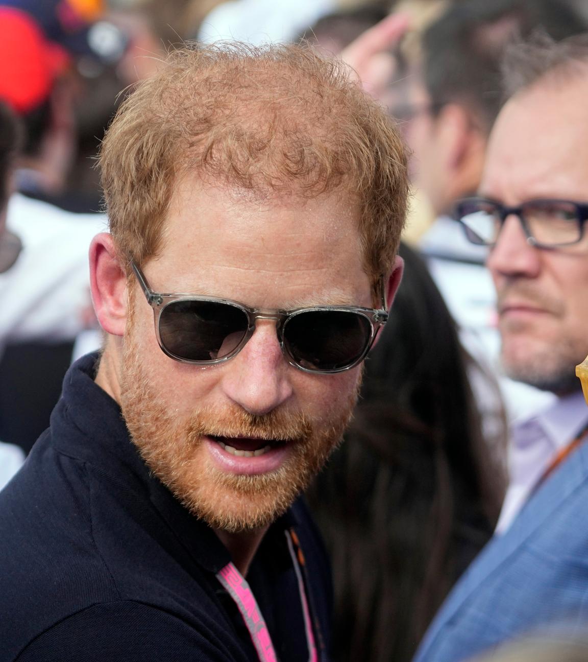 Prinz Harry trägt eine Sonnenbrille und steht zwischen mehrerem Menschen.