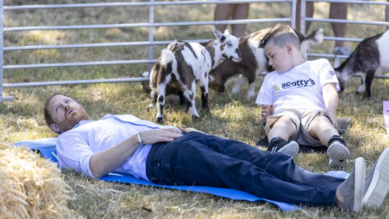 Prinz Harry liegt währen des Ziegenyogas auf einer Yogamatte. Neben ihm sitzt ein Kind und im Hintergrund stehen Ziegen. 