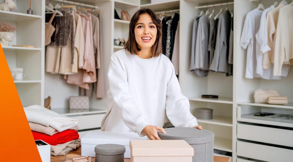 Frau packt Kisten für den Onlineverkauf ihrer Kleidung in einem begehbaren Kleiderschrank