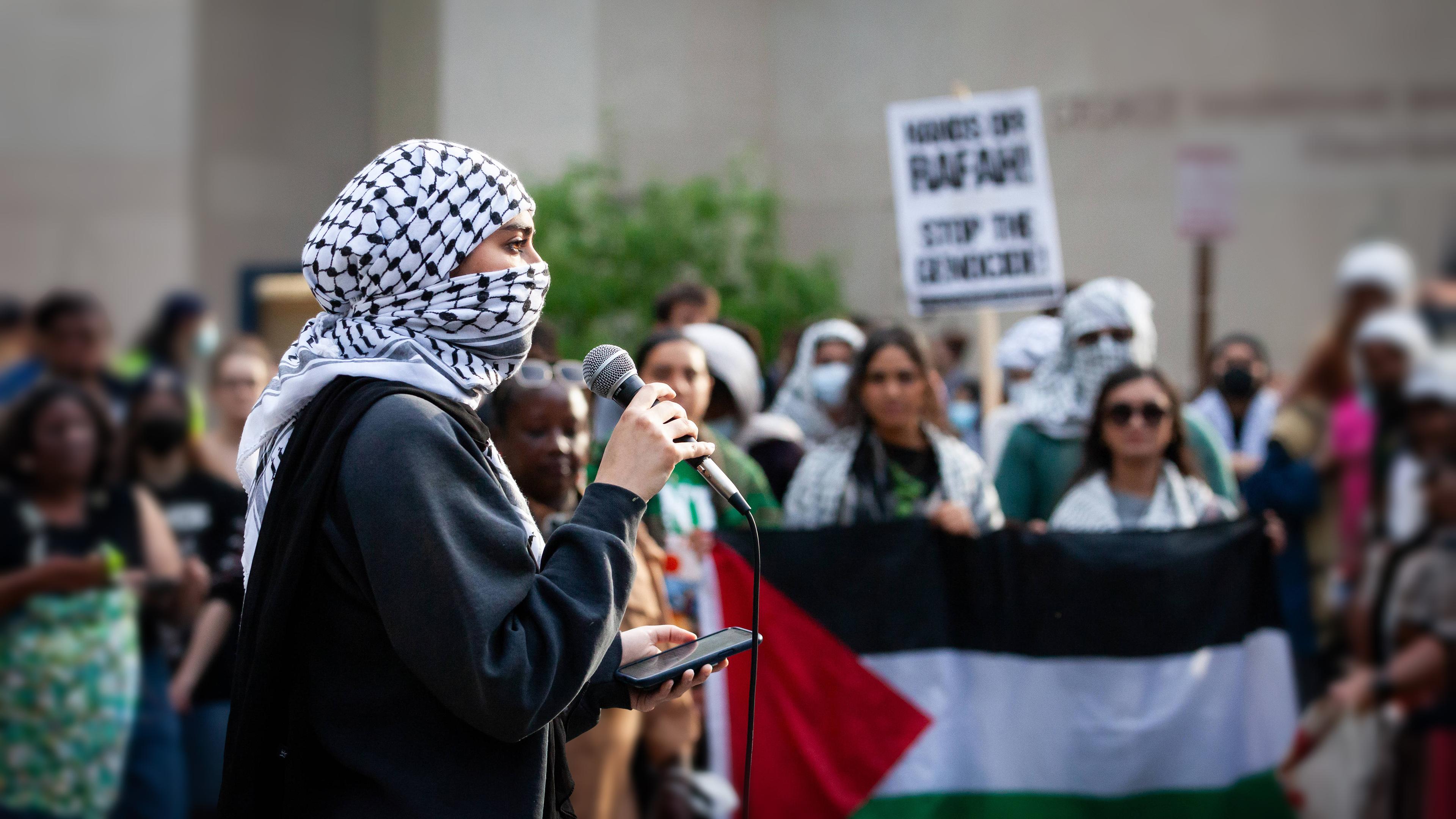 Eine mit einem Palästinensertuch vermummte junge Frau spricht in ein Mikrofon, im Hintergrund sind Demonstranten zu sehen.