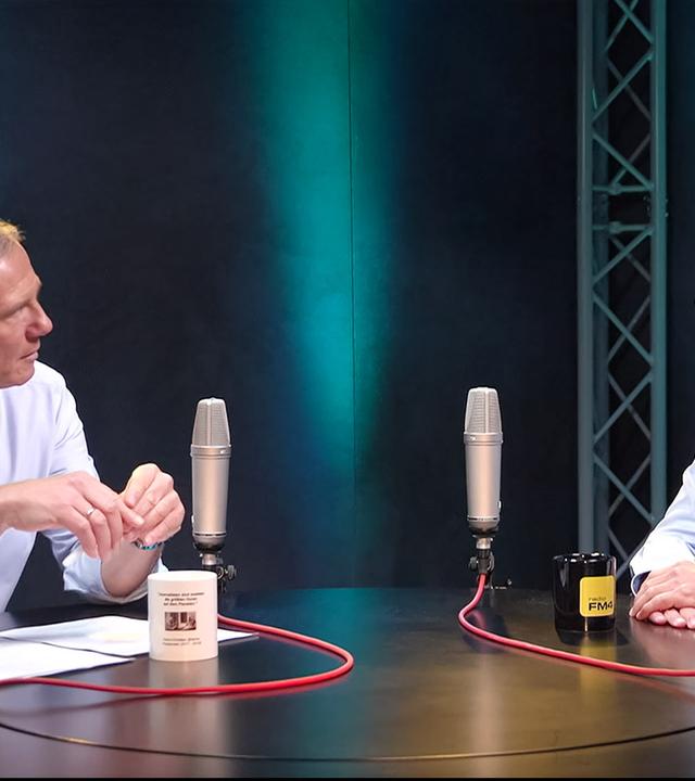 Das Bild zeigt zwei Männer, die an einem runden Tisch in einem Studio sitzen. Die Männer sind in formeller Kleidung gekleidet: der linke trägt ein weißes Hemd mit einem lila Krawatte, und der rechte ein hellblaues Hemd mit einem gemusterten, hellblauen Schlips.   Vor ihnen befinden sich zwei Mikrofone und ein schwarzer Becher mit dem Aufdruck „FM4“. Auf dem Tisch liegt ein Blatt Papier. Die Hintergrundbeleuchtung des Studios ist in sanften Blautönen gehalten. Der Raum hat eine minimalistische Gestaltung und vermittelt eine professionelle Atmosphäre.   Im Kontext des Bildes wird auf eine Sendung hingewiesen, die sich mit dem politischen System in Österreich beschäftigt.