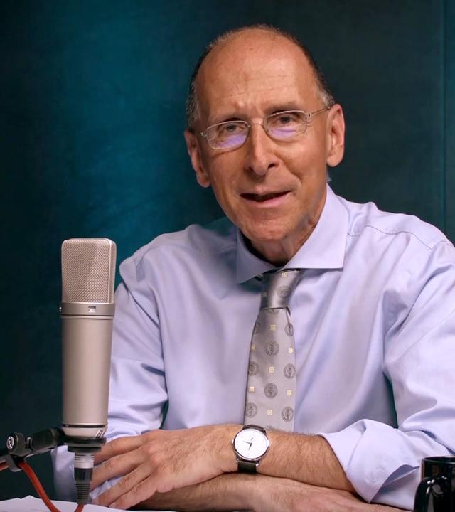 Das Bild zeigt einen Mann mittleren Alters, der an einem Tisch sitzt. Er trägt ein hellblaues Hemd und eine graue Krawatte mit einem Muster. Auf seinem Handgelenk befindet sich eine Armbanduhr. Vor ihm stehen ein Mikrofon, ein Blatt Papier und eine schwarze Tasse. Der Hintergrund des Bildes ist in sanften Blau- und Grüntönen gehalten. Der Mann lächelt und sieht direkt in die Kamera. Es scheint sich um eine Aufnahme für eine Fernsehsendung zu handeln, die möglicherweise politische Themen behandelt.