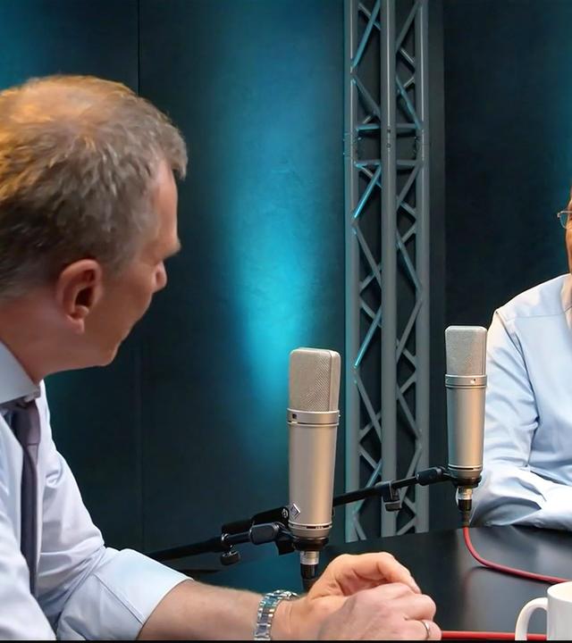 Die Abbildung zeigt zwei Männer, die in einem Studio miteinander sprechen. Es ist eine Multimedia-Sendung zu sehen, die sich mit dem politischen System in Österreich beschäftigt.    Im Vordergrund stehen zwei Mikrofone auf einem schwarzen Tisch. Der Mann links hat kurzes, graues Haar und trägt ein helles Hemd sowie eine Krawatte in dunklerer Farbe. Er schaut den anderen Mann an, dessen Gesicht von der Seite sichtbar ist.   Der Mann rechts hat eine Glatze und trägt eine Brille. Er hat ein hellblaues Hemd an und eine gemusterte Krawatte. Vor ihm steht eine schwarze Tasse. Die Wände des Studios sind dunkel und mit blauen Lichtakzenten beleuchtet, die im Hintergrund sichtbar sind.   Im Bild ist eine entspannte Atmosphäre zu erkennen, die auf ein informatives Gespräch hindeutet.