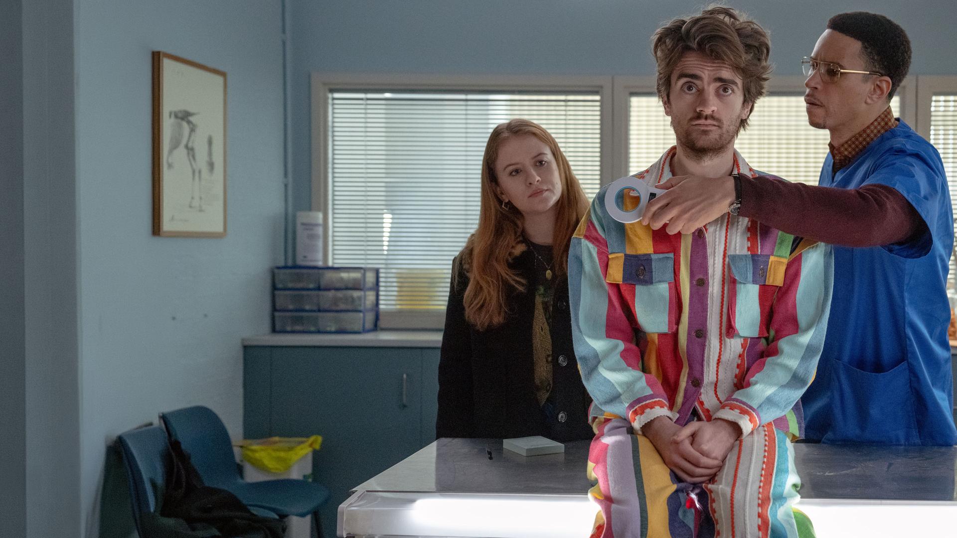 "Extraordinary - Projekt Haustier": In einem Arztzimmer sitzt Jizzlord (Luke Rollason) auf einem Behandlungstisch und blickt skeptisch geradeaus. Hinter ihm steht Dr. Wenderburn (John Macmillan), der einen Metall-Detektor vor seine Brust hält. Jen (Máiréad Tyers) steht ebenfalls hinter Jizzlord und betrachtet ihn nachdenklich.