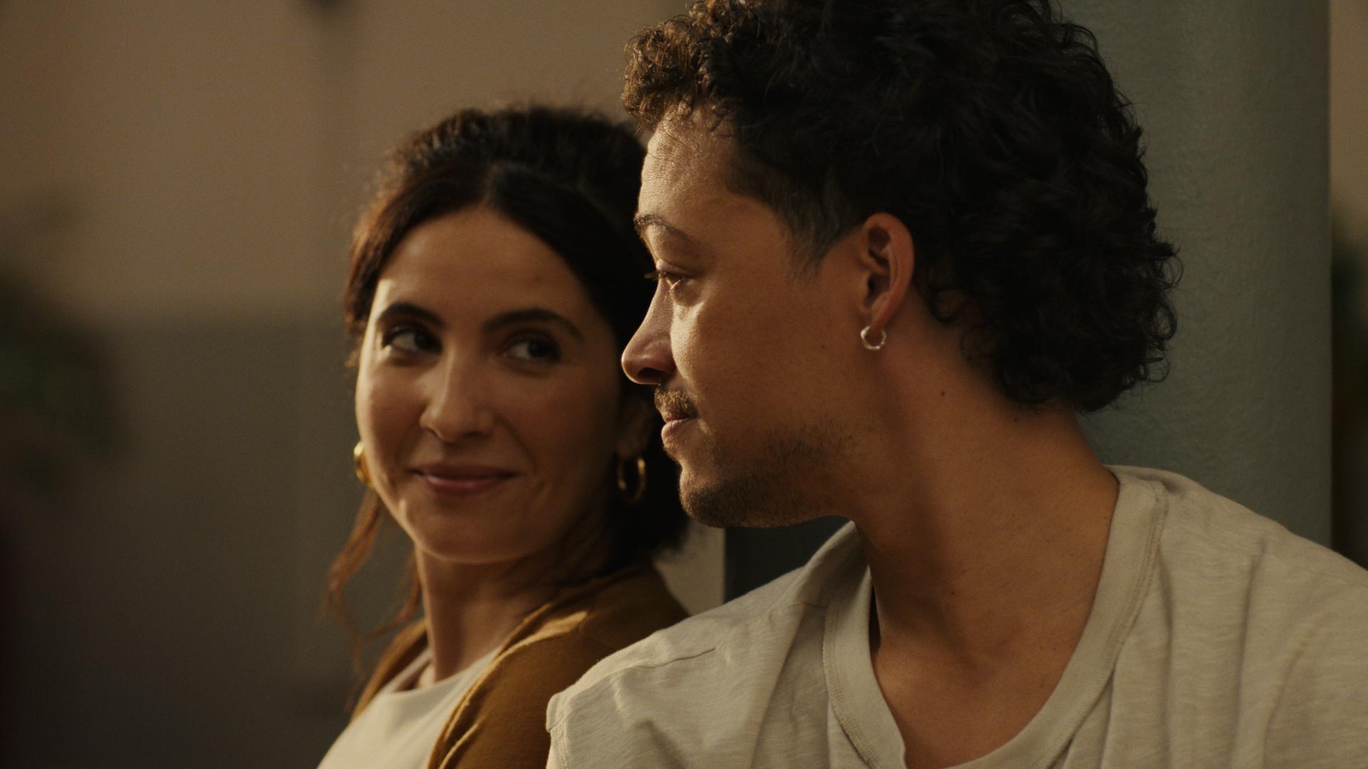 "The other gAIrl – prompt: verknallt": Nahaufnahme: Lisa (Chryssanthi Kavazi) und Rafa (Aurel Mertz) sitzen dicht nebeneinander. Lisa lächelt verlegen, während Rafa sie aufmerksam ansieht.