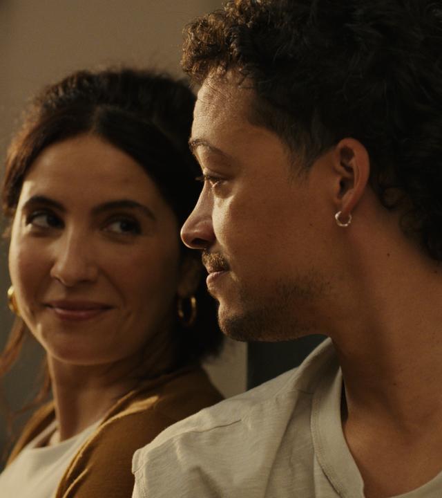 "The other gAIrl – prompt: verknallt": Nahaufnahme: Lisa (Chryssanthi Kavazi) und Rafa (Aurel Mertz) sitzen dicht nebeneinander. Lisa lächelt verlegen, während Rafa sie aufmerksam ansieht.