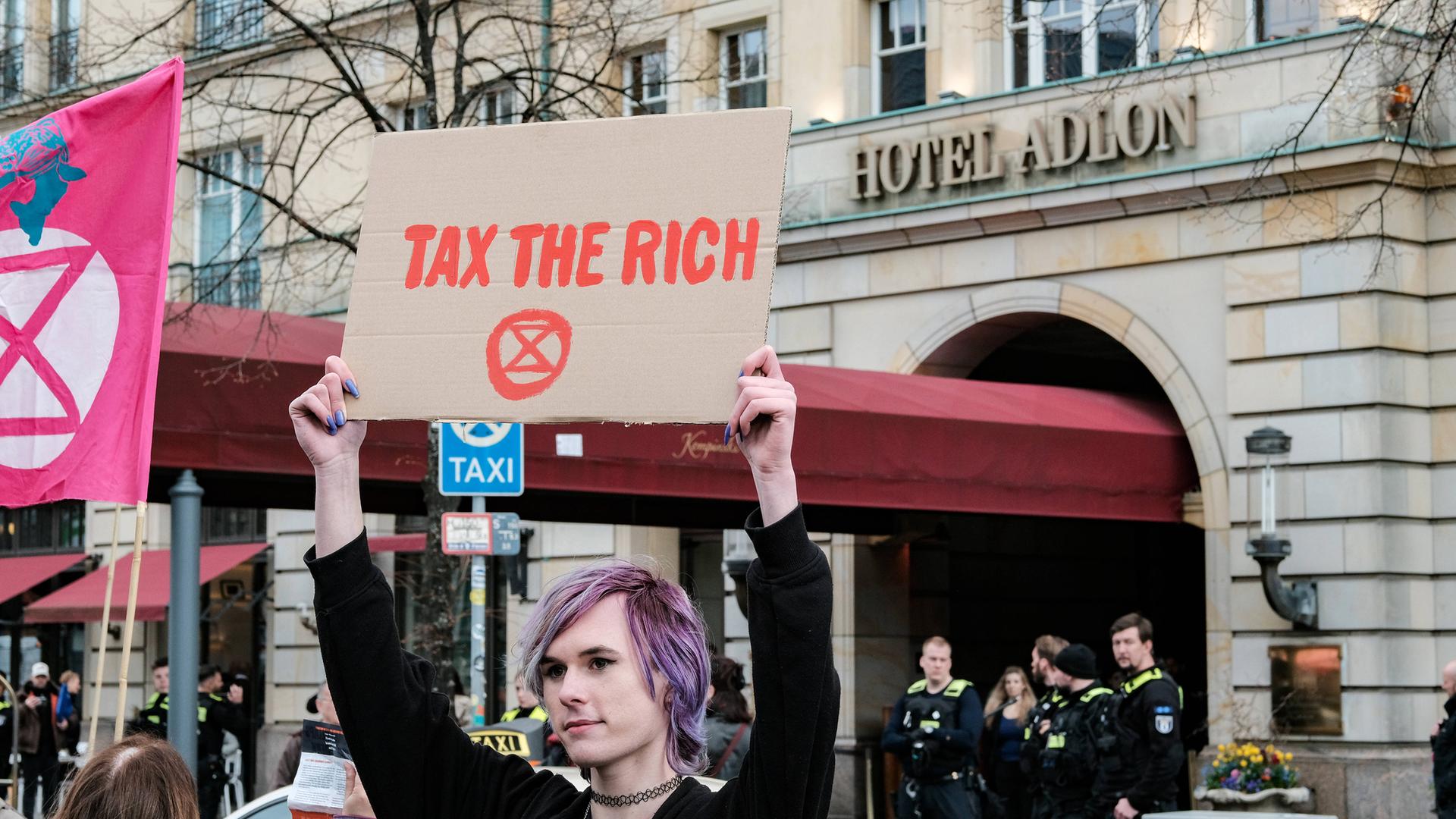 Eine junge Frau mit lila gefärbten Haaren hält bei einer Demonstration vor dem Luxushotel Adlon in Berlin ein Schild hoch, auf dem tax the rich steht.