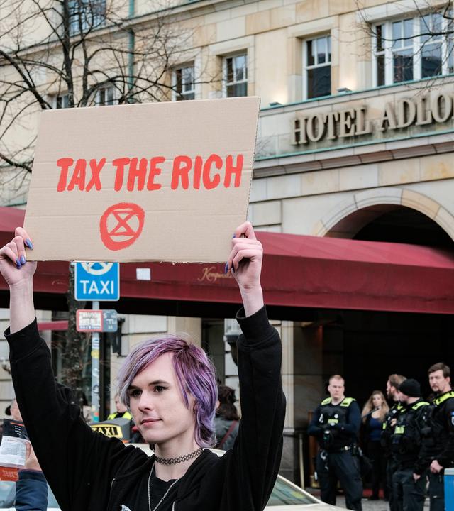 Eine junge Frau mit lila gefärbten Haaren hält bei einer Demonstration vor dem Luxushotel Adlon in Berlin ein Schild hoch, auf dem tax the rich steht.