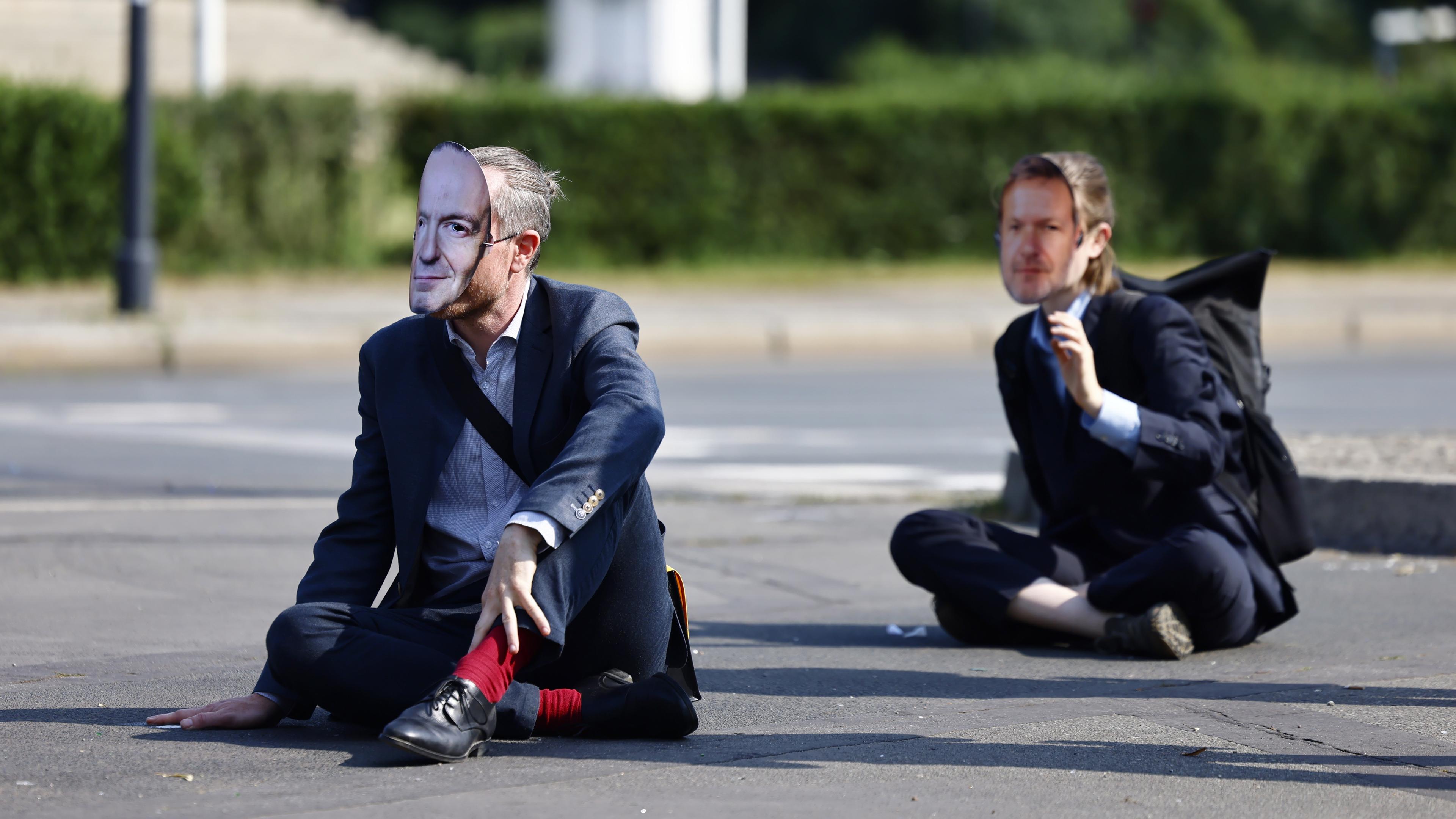 Zwei Menschen mit Pappmasken sitzen auf einer Straße.