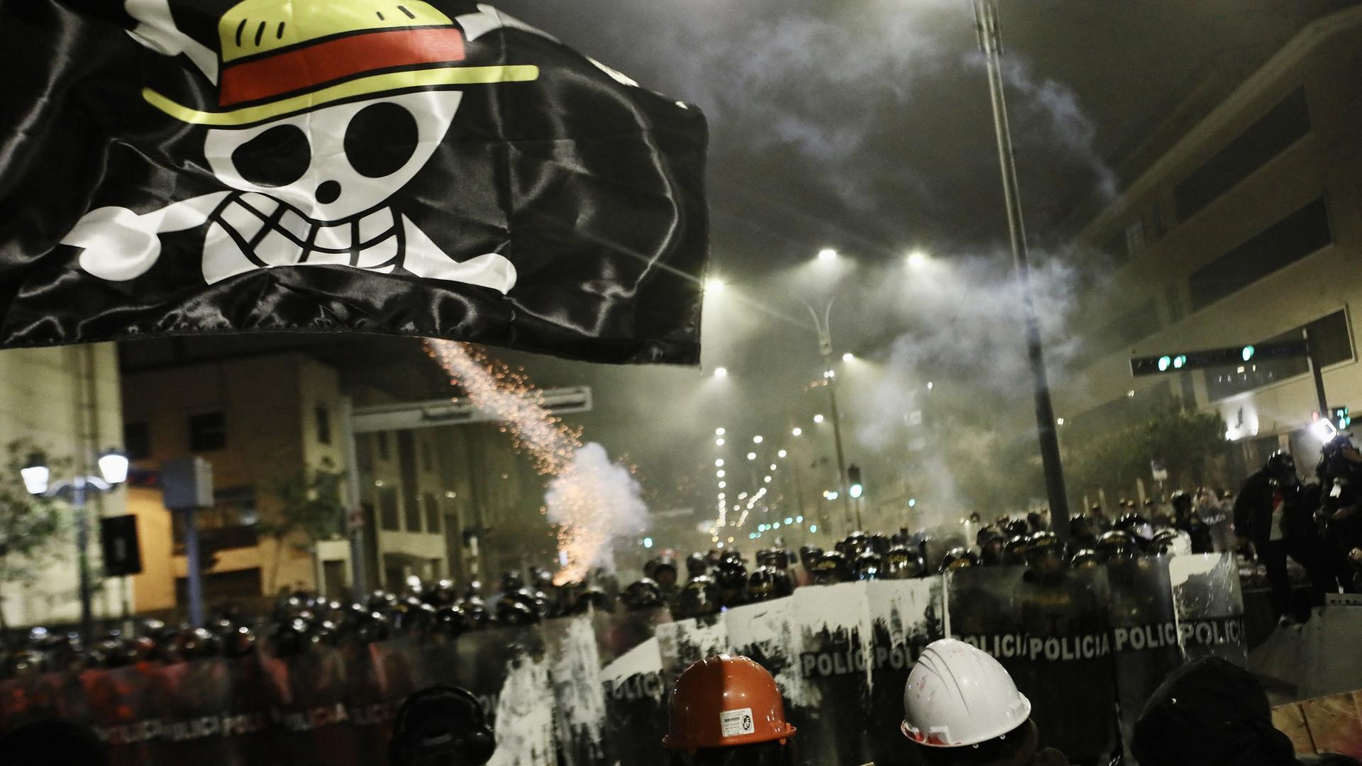 Demonstration bei Nacht: Polizisten mit Schilden sperren Demonstranten den Weg ab. Die Nacht wird von Straßenlampen und Feuerwerkskörpern erleuchtet. Über allem schwebt eine scharze Flagge mit einer Skelett-Figur, die einen Strohhut trägt