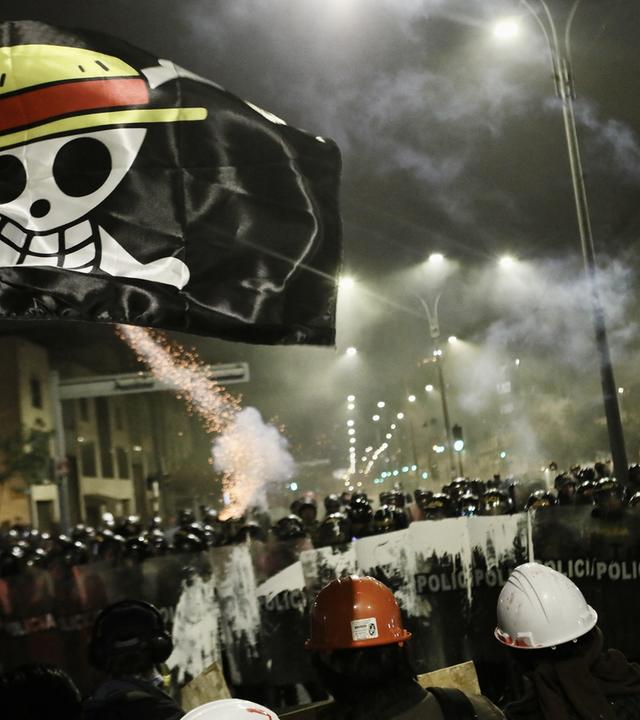 Demonstration bei Nacht: Polizisten mit Schilden sperren Demonstranten den Weg ab. Die Nacht wird von Straßenlampen und Feuerwerkskörpern erleuchtet. Über allem schwebt eine scharze Flagge mit einer Skelett-Figur, die einen Strohhut trägt