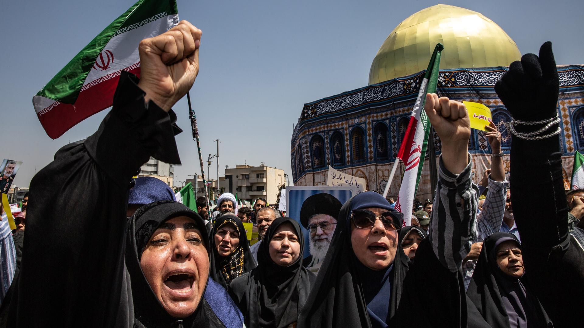 Frau ruft mit erhobener Faust vor iranischer Flagge, im Hintergrund eine Moschee.