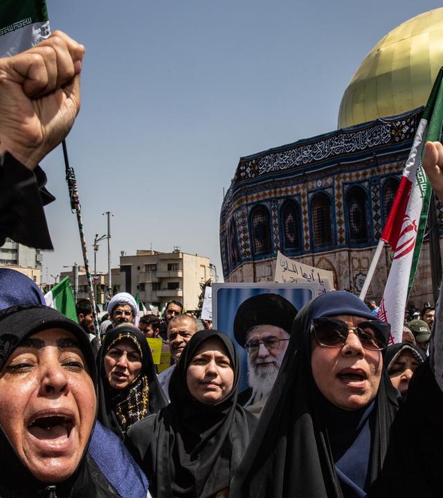 Frau ruft mit erhobener Faust vor iranischer Flagge, im Hintergrund eine Moschee.