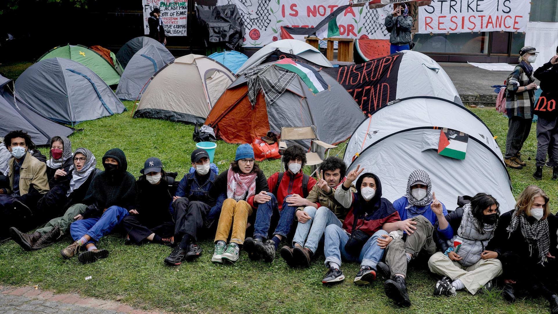 Eine Reihe junger Menschen mit Gesichtsmasken sitzen nebeneinander mit verschränkten Armen auf dem Rasen. Hinter ihnen sind Zelte aufgebaut und Plakate mit Slogans gegen den Krieg in Gaza aufgestellt