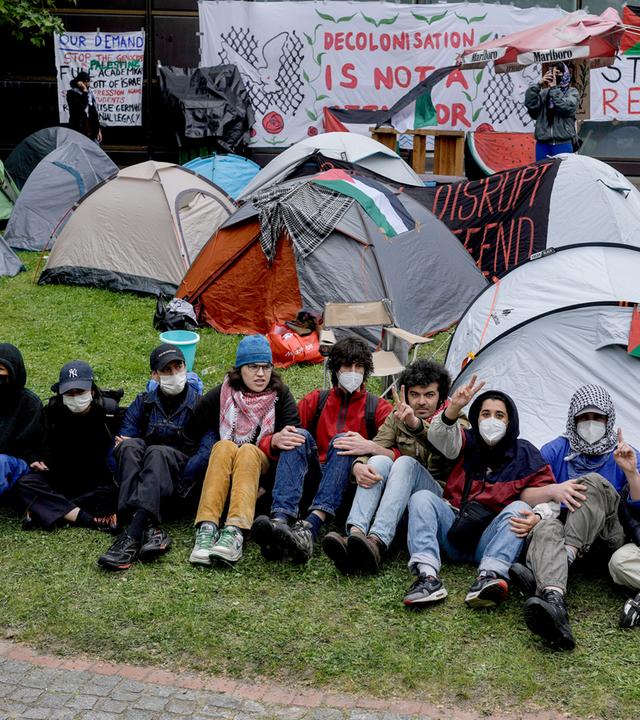 Eine Reihe junger Menschen mit Gesichtsmasken sitzen nebeneinander mit verschränkten Armen auf dem Rasen. Hinter ihnen sind Zelte aufgebaut und Plakate mit Slogans gegen den Krieg in Gaza aufgestellt