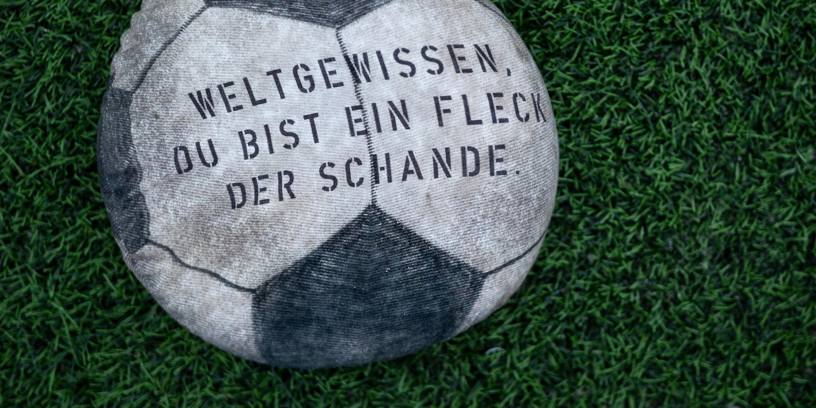 Ein mit Sand gefüllter Stoff-Fußball, auf dem "Weltgewissen, du bist ein Fleck der Schande" steht, liegt auf dem Rasen des Fußballstadions am Schloss Strünkede in Herne.
